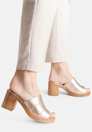 Metaliczne złote mule o błyszczącej powierzchni, z otwartym czubkiem i blokowym, drewnianym obcasem, zestawione z jasnymi, dopasowanymi spodniami.