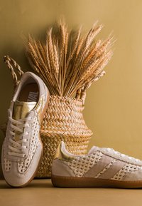 Panier tressé avec des tiges de blé séchées derrière une paire de baskets blanches tissées avec des accents dorés sur un fond beige.