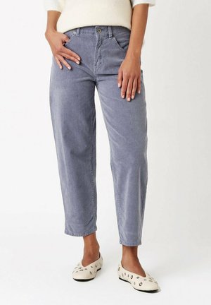 Woman wearing light purple corduroy straight-leg pants with front pockets and cream flats with black polka dots, standing against white background.