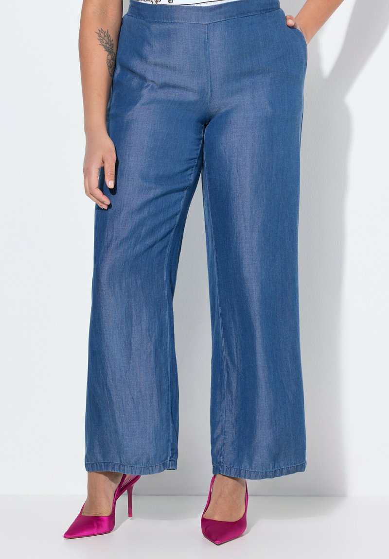Dżinsowe spodnie z szerokimi nogawkami o gładkiej fakturze, średnim stanem, dwoma bocznymi kieszeniami i widocznym przeszyciem. Zestawione z różowymi wysokimi obcasami.