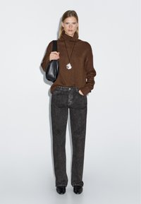 Maglione marrone con collo alto, abbinato a jeans dritti grigio scuro. La modella tiene una borsa nera e indossa una collana con pendente.