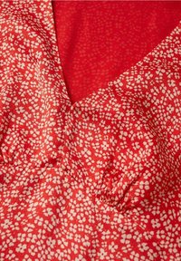 Roter, floral gemusterter Stoff mit kleinen weißen Blumen. Das Material scheint weich zu sein, mit gerafften Details am Ausschnitt und einer strukturierten Hintergrundfläche.