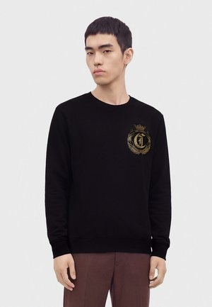 Junger Mann mit kurzen schwarzen Haaren trägt schwarzen Pullover mit goldenem Emblem auf der Brust und braune Hose, steht vor einfarbigem Hintergrund.