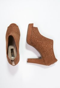 Zapatos de tacón alto de ante marrón con diseño perforado, abertura en los dedos y acento de cremallera metálica en el tacón.