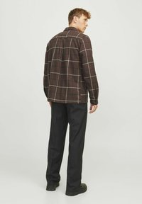 Bruine geruite shirtjas met een gestructureerde kraag, lange mouwen en contrasterende witte lijnen; gecombineerd met zwarte broek en zwarte schoenen.