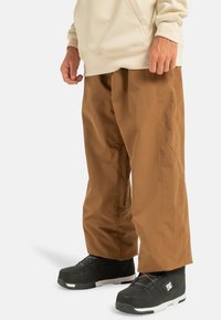 Bruine, losse broeken van een gladde stof, gecombineerd met zwarte schoenen met witte accenten en een gestructureerde zool.