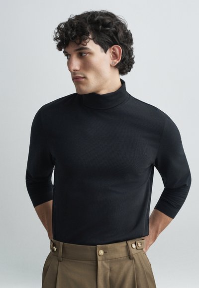 Maglione nero a collo alto con maniche a tre quarti, realizzato in un materiale liscio, abbinato a pantaloni beige a vita alta con bottoni dorati.