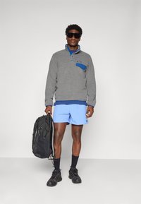 Man in een grijze fleece trui, lichte blauwe shorts, zwarte zonnebril, zwarte wandelschoenen, met een zwarte rugzak tegen een effen achtergrond.
