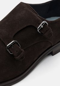 Chaussure en daim marron à la forme arrondie, dotée de deux boucles en argent et d'une couture contrastante le long des bords. Doublure intérieure lisse.