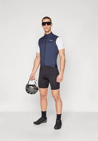 Strój kolarski z dopasowaną granatową kamizelką i czarnymi szortami, wykonany z oddychającej tkaniny odprowadzającej wilgoć. Model trzyma czarny kask, ma na sobie ciemne okulary przeciwsłoneczne.