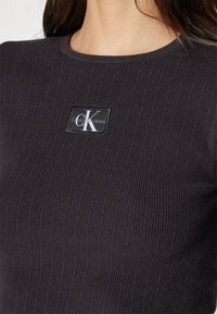 Sort ribbet top med rund halsudskæring, prydet med et rektangulært logo-patch med "Calvin Klein Jeans" i hvid og sort.