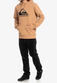Felpa con cappuccio color sabbia con logo di montagna nero e testo sulla parte anteriore, abbinata a pantaloni in corduroy neri e scarpe nere. Presenta una tasca a marsupio.