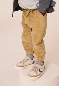 Beige corduroy-housut, joissa on väljä istuvuus ja elastiset hihnat, yhdistettynä harmaisiin ja beigeihin lenkkareihin, joissa on sinisiä ja ruskeita yksityiskohtia.