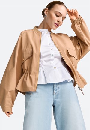 Junge Frau in einer beigen, übergroßen Jacke, weißer, geknöpfter Bluse und hellblauen Jeans, die mit geneigtem Kopf und erhobener Hand nahe der Stirn posiert.