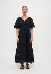 Robe maxi noire avec des manches courtes et transparentes, devant à boutons et accents en dentelle au niveau de l'ourlet. Fabriquée en tissu léger avec un design à volants.