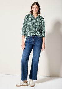 Blusa verde a fantasia con maniche 3/4 e scollo a V, abbinata a jeans a gamba larga blu scuro e sneaker beige. Texture del tessuto morbida.