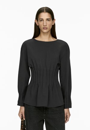 Schwarzes Langarmoberteil mit weitem Ausschnitt, gerafftem strukturiertem Design in der Taille und leicht ausgestelltem Saum. Kombiniert mit dunklen Jeans.