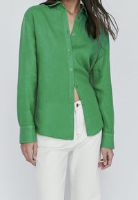Woman wearing a bright green long-sleeve button-up shirt partially tucked into high-waisted white pants against a plain background.