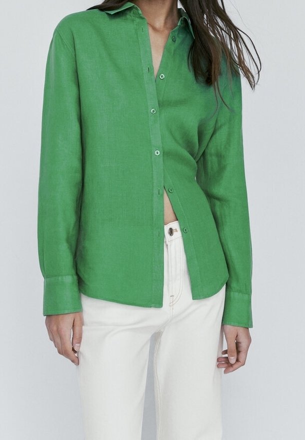 Woman wearing a bright green long-sleeve button-up shirt partially tucked into high-waisted white pants against a plain background.