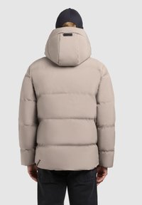 Puffer jacket in light beige, featuring a high collar, attached hood, and quilted texture. Dark pants visible below the jacket.