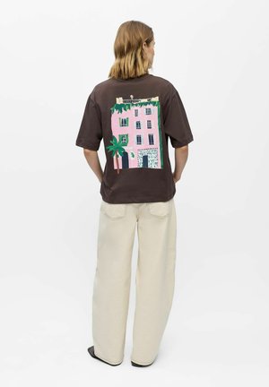 Person med skulderlangt hår iført mørkebrun T-shirt med pink bygning og grøn palme på ryggen, cremefarvede vide bukser, sorte sko.