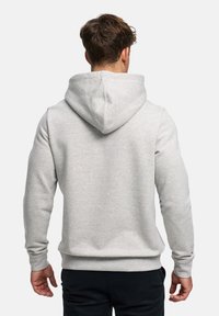 Man met kort bruin haar, gekleed in een lichtgrijze hoodie en zwarte broek, gezien van achteren tegen een effen witte achtergrond.