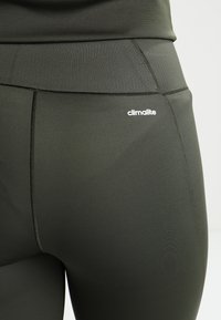 Ciemnozielone legginsy sportowe o gładkiej strukturze, z wysokim stanem i białym logo "climalite" blisko górnego szwu.