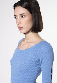 Top azul acanalado con escote redondeado y mangas largas. Detalles notables: puños abiertos adornados con tres botones decorativos. Textura suave.