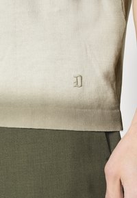 Pletený top v světle béžové barvě s přechodem do olivově zelené. Má malé, embosované logo "D" na lemu. Hladká textura, přiléhavý design.