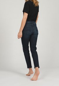 Tmavě modré džíny s úzkým střihem, středně vysokým pasem a zadními kapsami, kombinované s černým topem s krátkým rukávem. Stojící na boso.
