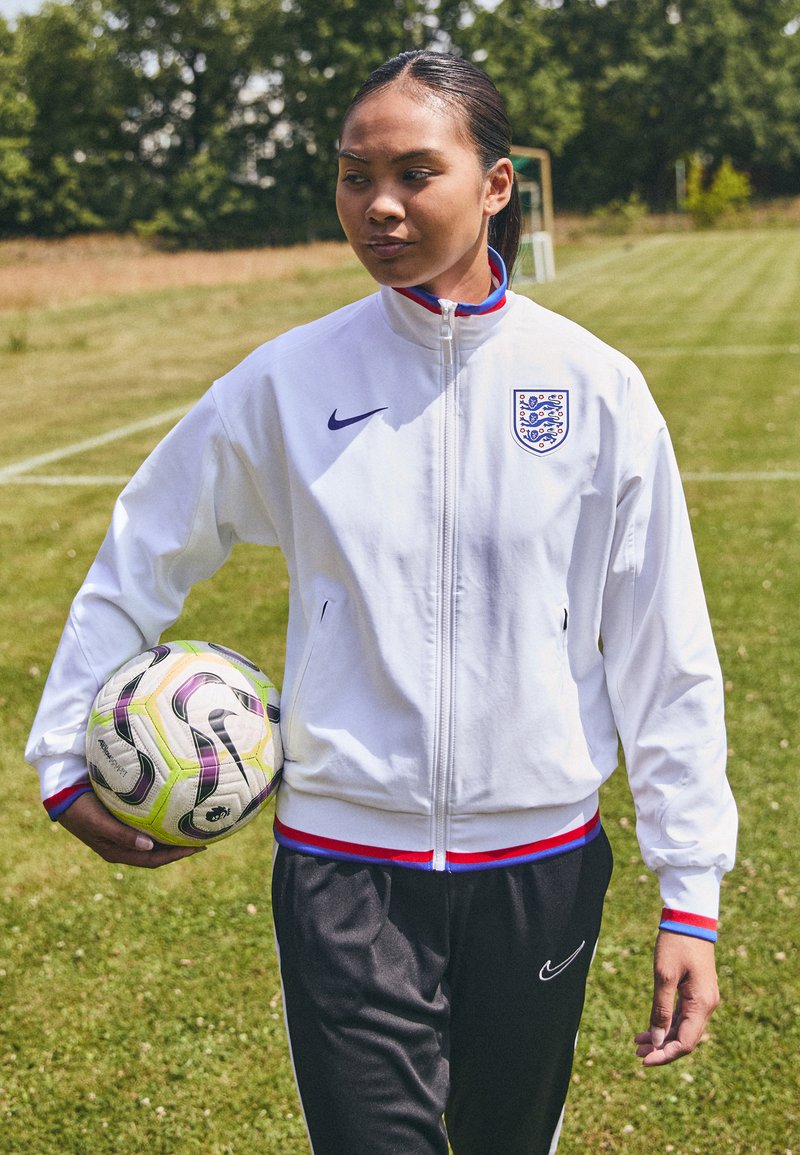 Hvit zip-up jakke med blå og røde detaljer, med England-merket, kombinert med sorte bukser og holder en flerfarget fotball.