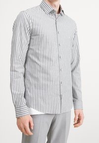 Camisa de botones a rayas en gris claro con rayas verticales en gris oscuro. Cuenta con un collar, mangas largas y un dobladillo curvado.