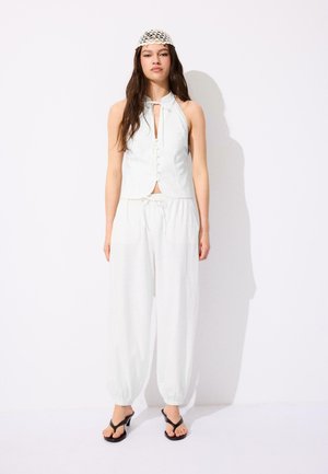 Mlada žena s dugom tamnom kosom nosi bijelu majicu bez rukava s gumbima, bijele hlače s vezicom, crne sandale na povišenoj peti i bijelu kukičanu kapu.