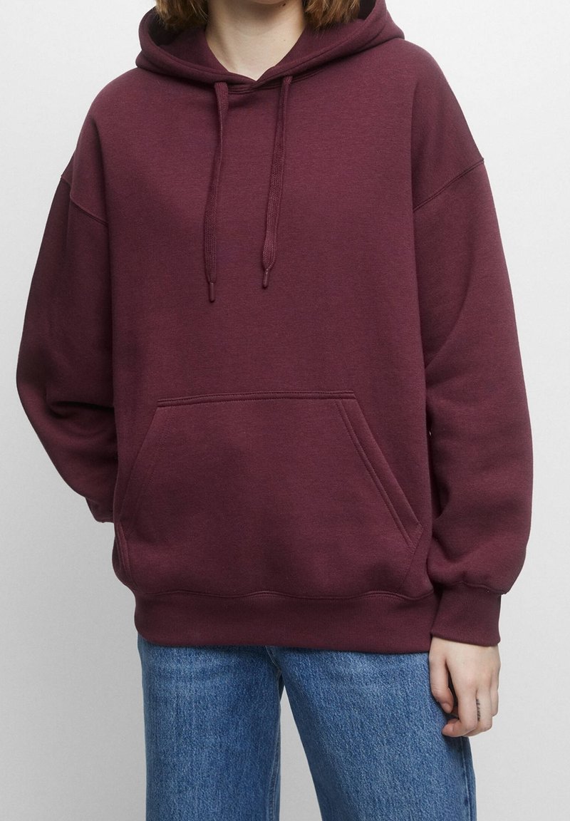 Personne portant un sweat à capuche bordeaux avec poche ventrale et un jean bleu, vue des épaules aux hanches sur un fond uni.