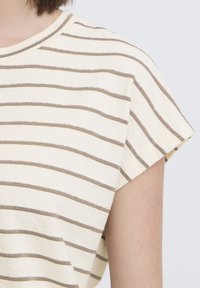 Gros plan sur une chemise à manches courtes beige avec de fines rayures horizontales brunes portée par une personne devant un fond clair uni.