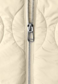 Primo piano di una cerniera in metallo argentato su un tessuto trapuntato color crema, che mostra i motivi di cucitura e il dettaglio della linguetta della cerniera.