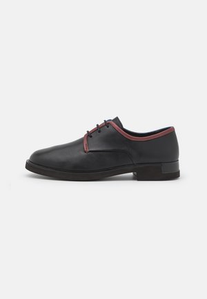 Chaussure habillée en cuir noir avec bordure marron, trois œillets et petit talon, présentée de profil sur fond blanc.