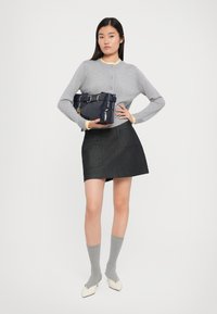 Cardigan grigio lavorato a maglia con bottoni, mini gonna nera, calze grigie fino al ginocchio e décolleté bianchi a punta. Tiene in mano una borsa blu navy con dettaglio a cintura.