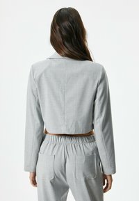 Chaqueta gris cortada con mangas largas y cuello con muesca, hecha de una tela con una sutil textura. Combinada con pantalones grises a juego.