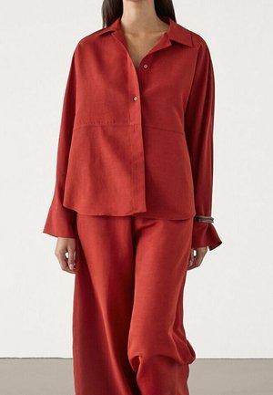 Femme portant une chemise rouge ample à manches longues avec un pantalon large assorti sur un fond uni.