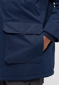 Chaqueta aislante de color azul marino que cuenta con un gran bolsillo frontal, tejido texturizado y detalle de puño ajustado, ofreciendo funcionalidad y calidez.
