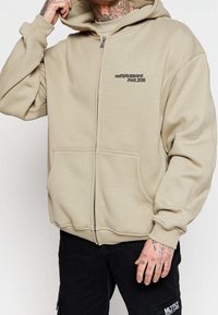 Beige mikina s kapucí na zip vyrobená z měkké tkaniny. Obsahuje klokaní kapsu, žebrované manžety a potištěné logo na hrudi.