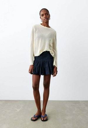 Frau trägt einen hellbeigen Pullover, schwarzen gestuften Minirock und blaue Flip-Flops, steht an einer schlichten weißen Wand und einem Betonboden.