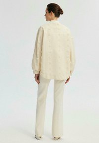 Veste surdimensionnée couleur crème avec motifs floraux texturés, épaules tombantes et ourlet arrondi, associée à un pantalon blanc ajusté.