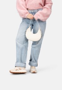 Sac à main en forme de croissant blanc tenu par une personne portant un pull en peluche rose et un jean bleu clair, associé à des baskets beiges.