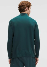 Vyras su trumpais raudonais plaukais, vilkintis tamsiai žalią ilgomis rankovėmis sportinį marškinėlį ir atitinkančias kelnes, atsukęs nugarą kamerai.