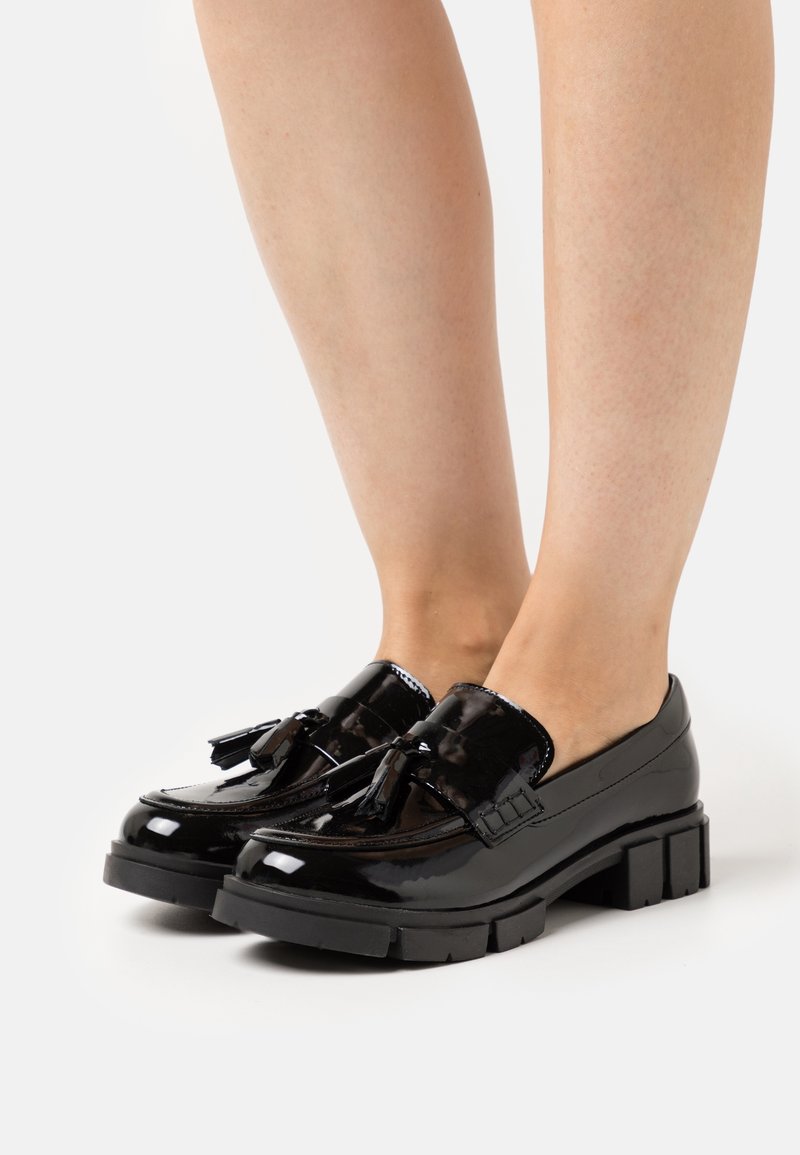 Mocassins en cuir verni noir avec des glands et des semelles épaisses portés pieds nus, présentés sur un fond blanc.
