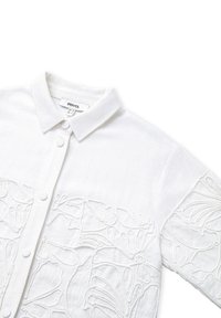 Camicia bianca in lino con colletto button-down e delicati ricami floreali sul petto e sulle maniche. Dettagli in tessuto testurizzato evidenti.