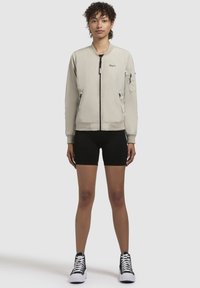 Geacă bomber de un gri deschis, cu fermoar frontal, buzunare laterale, manșete ribbate și logo brodat. Modelul poartă pantaloni scurți negri și adidași cu glezna înaltă.
