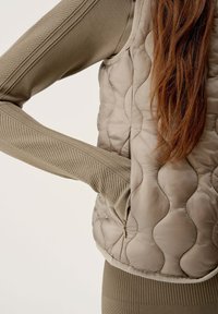 Gilet trapuntato beige con un tessuto a costine testurizzato sulle maniche. Presenta un collo alto e una cerniera laterale. Il materiale esterno ha una finitura morbida e lucida.